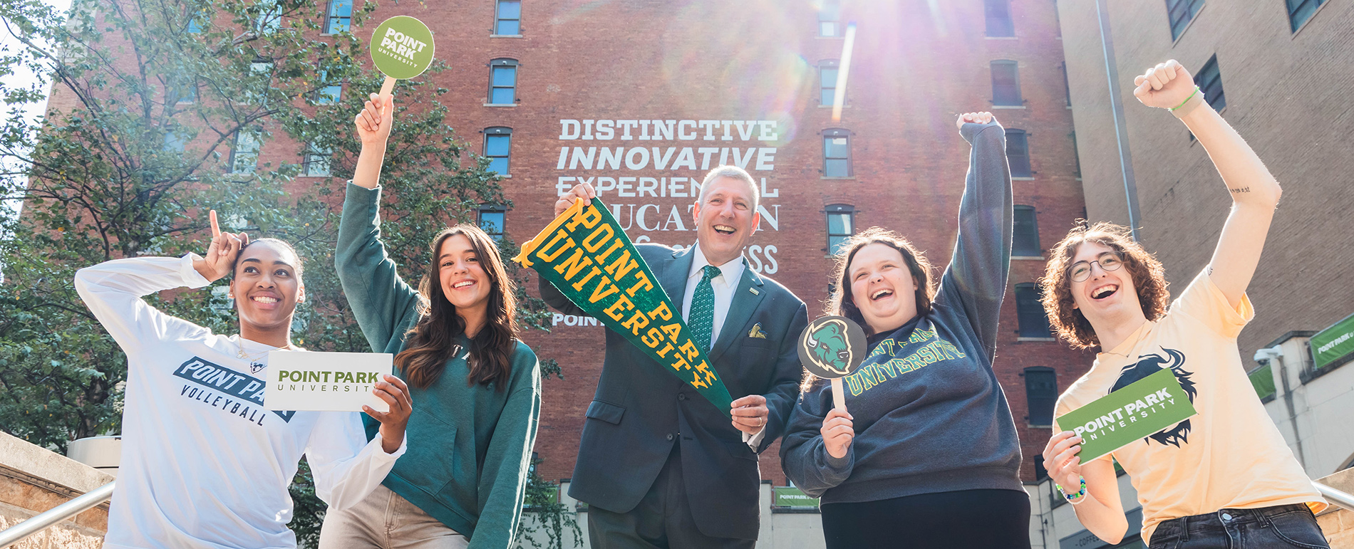 Students pose with President Brussalis in village park holding signs and pennants.