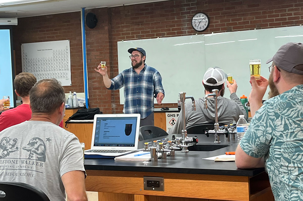 A man stands in a classroom lab holding up a small glass of fluid.