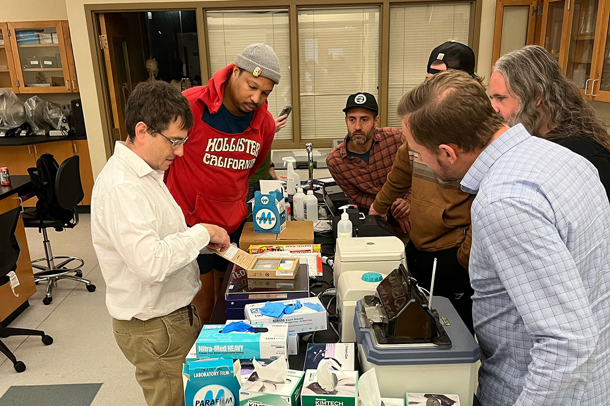 A group of people surrounds a teacher in a classroom lab.