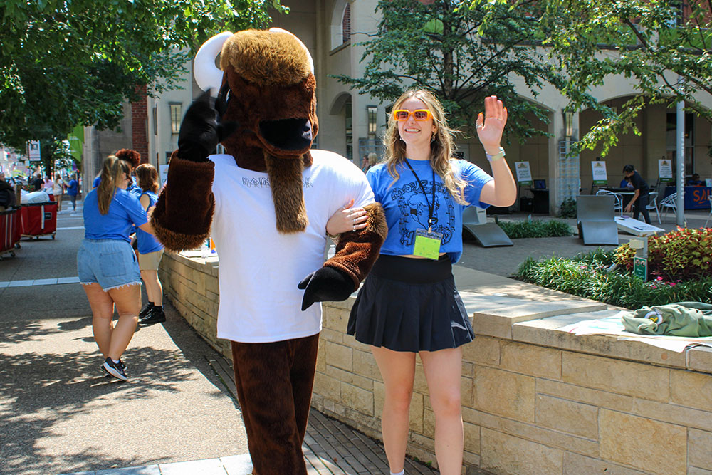 The bison mascot and a Pioneer Ambassador link arms and wave at the camera.