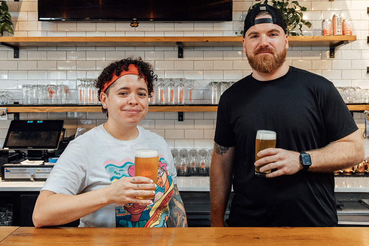 A woman and man stand behind a bar holding two glasses of beer.