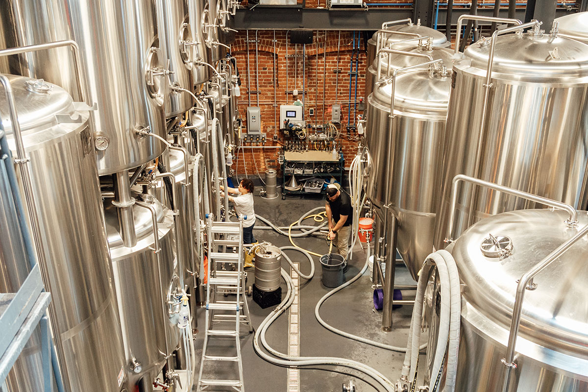 Two people work in a brewery, surrounded by silver tanks.