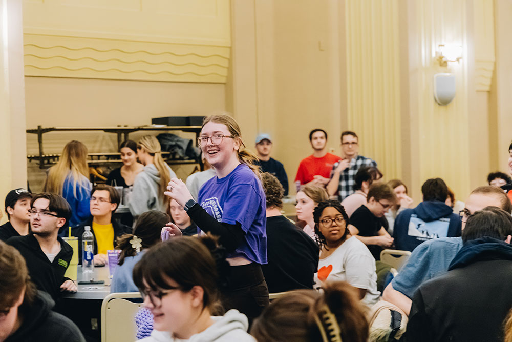 A student runs through a room of tables filled with students.