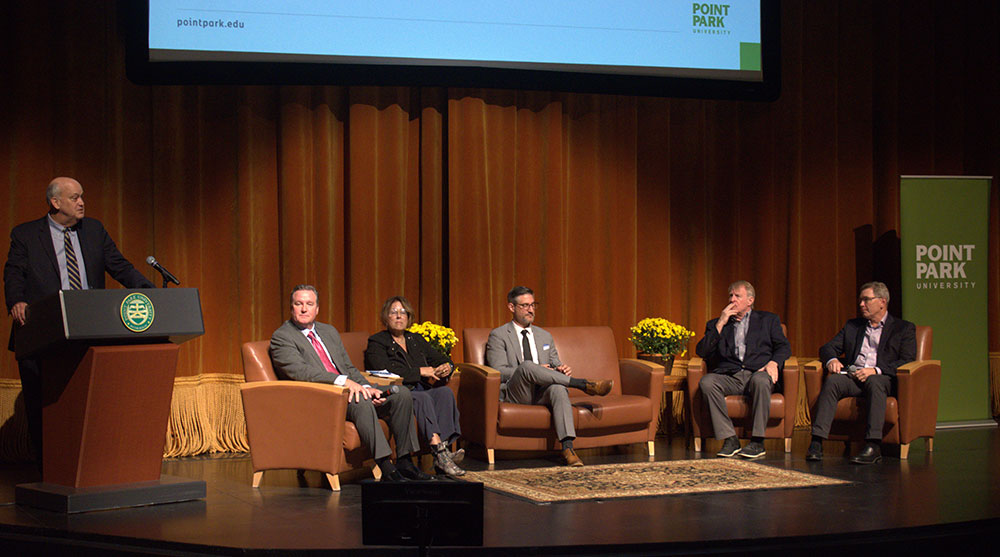 A man stands at a podium near 5 people sitting on couches and chairs on a stage.