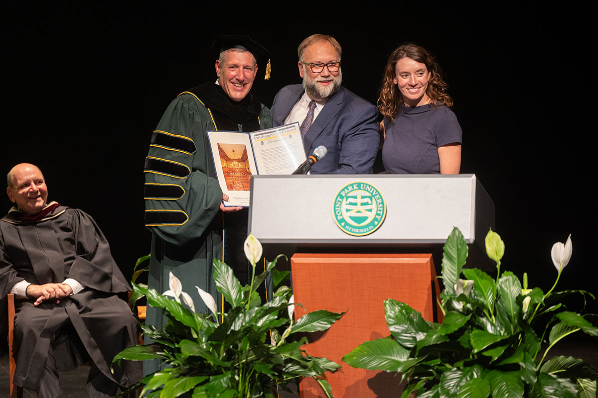 President Brussalis stands at the podium with a man and woman, holding a proclamation.