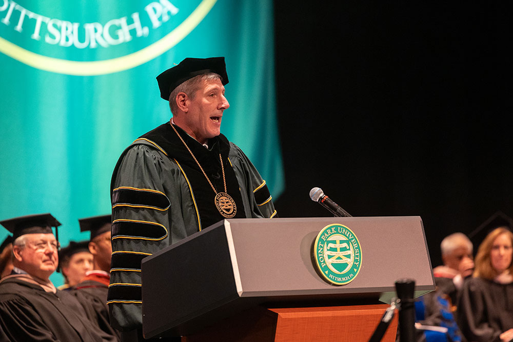 A man in green academic regalia speaks at a podium.
