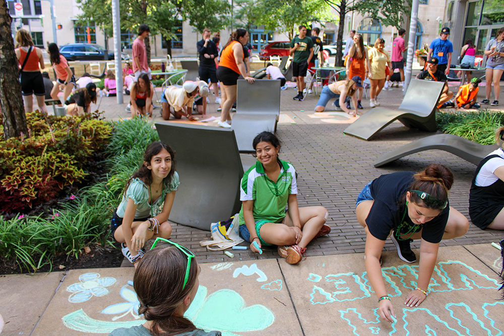 A large group of students sit on the sidewalk and draw welcome messages.